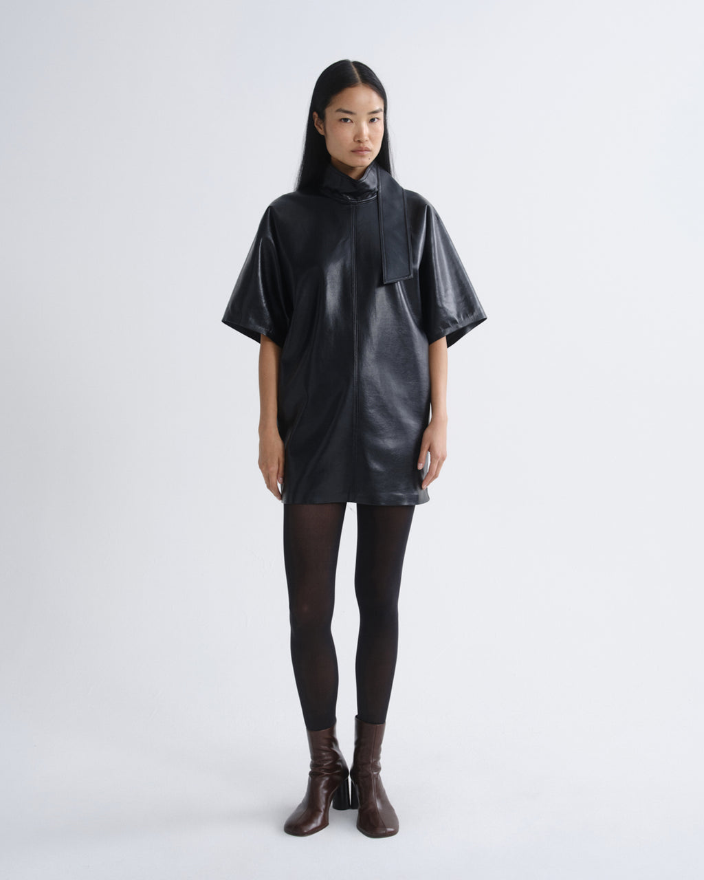 A model stands against a light gray studio background wearing a black faux-leather poncho-style top with a high collar and wide short sleeves, paired with black tights and dark brown heeled ankle boots.