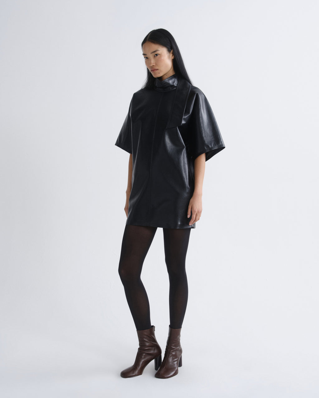 A model wearing a glossy black short-sleeved poncho-style top, sheer black tights, and dark brown heeled ankle boots, standing against a light gray studio background.