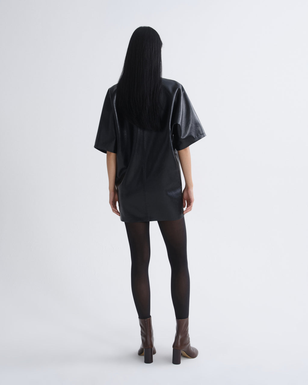 A model viewed from behind wearing a black oversized short-sleeve top with a glossy finish, paired with black tights and brown ankle boots, against a light gray studio background.