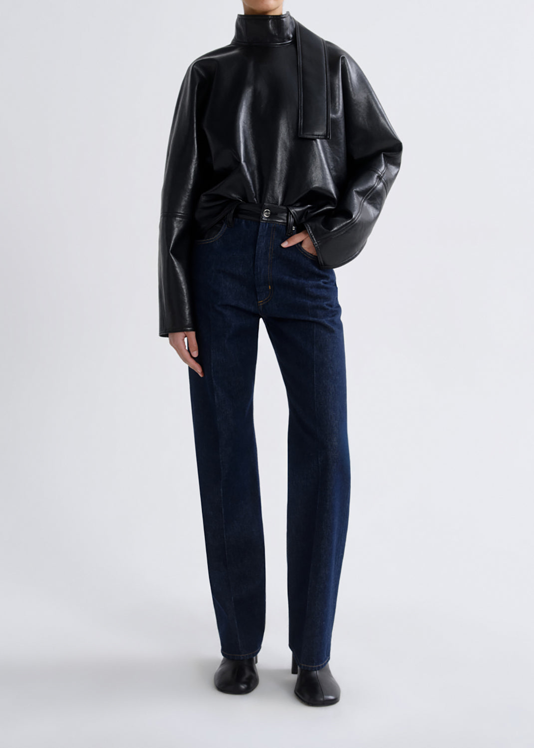 A model wearing a black faux-leather mock-neck jacket with wide sleeves and dark blue straight-leg jeans, standing against a grey background.