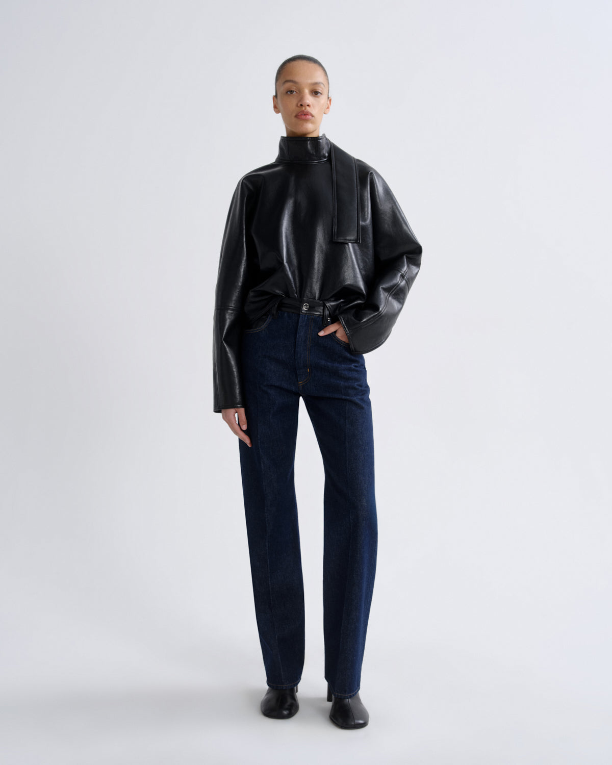 A model stands against a light gray studio background wearing a glossy black long-sleeve mock-neck top, dark blue straight-leg jeans, and black shoes.