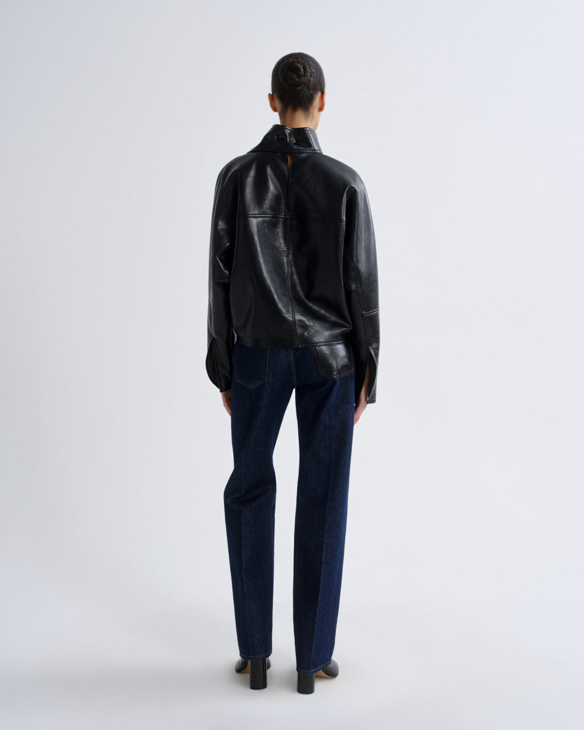 A model viewed from the back wearing a black leather jacket and dark blue jeans against a light gray studio background.