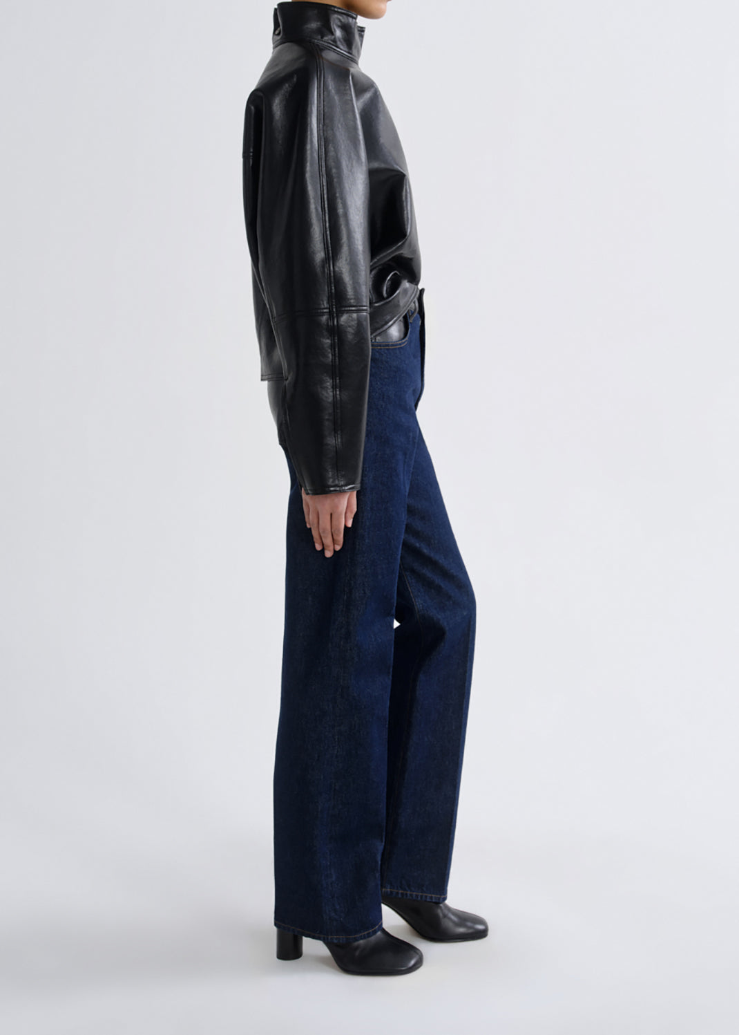 A model shown from the side wearing a black leather mock-neck jacket, dark blue wide-leg jeans, and black leather boots against a grey background.