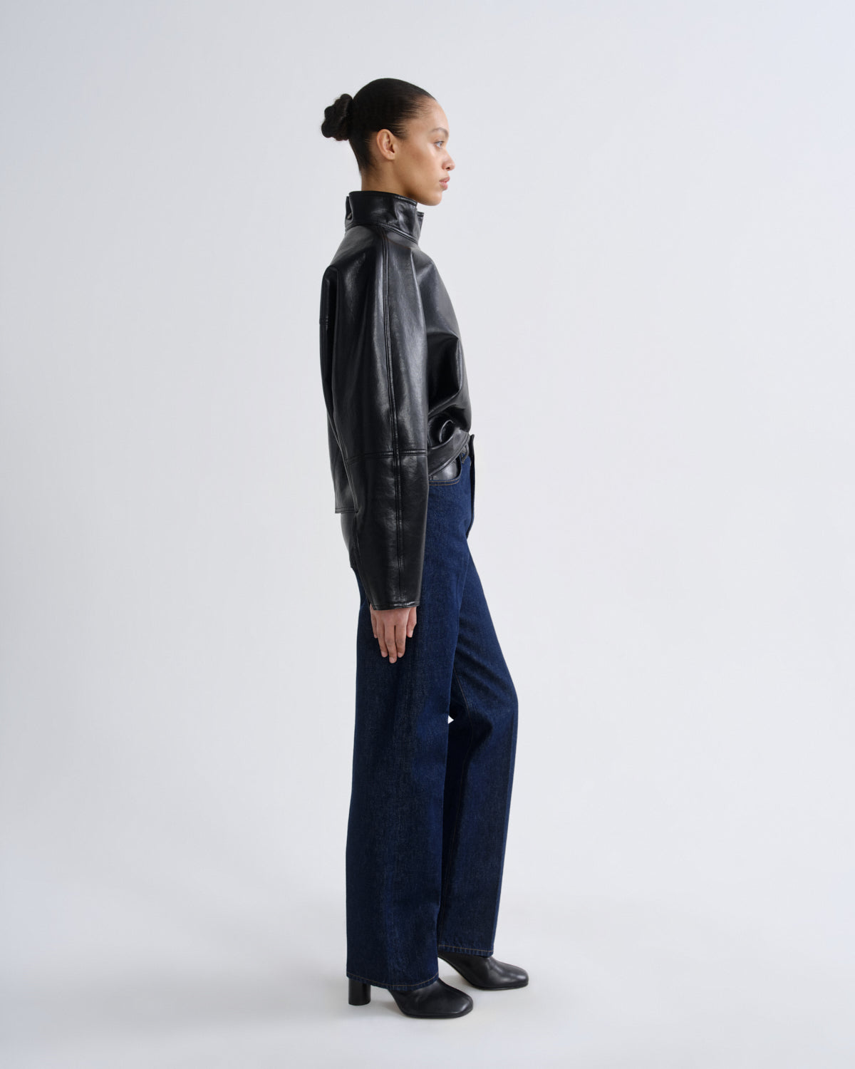 A model shown in profile wearing a black leather jacket, dark blue wide-leg jeans, and black shoes, standing against a light gray studio background.