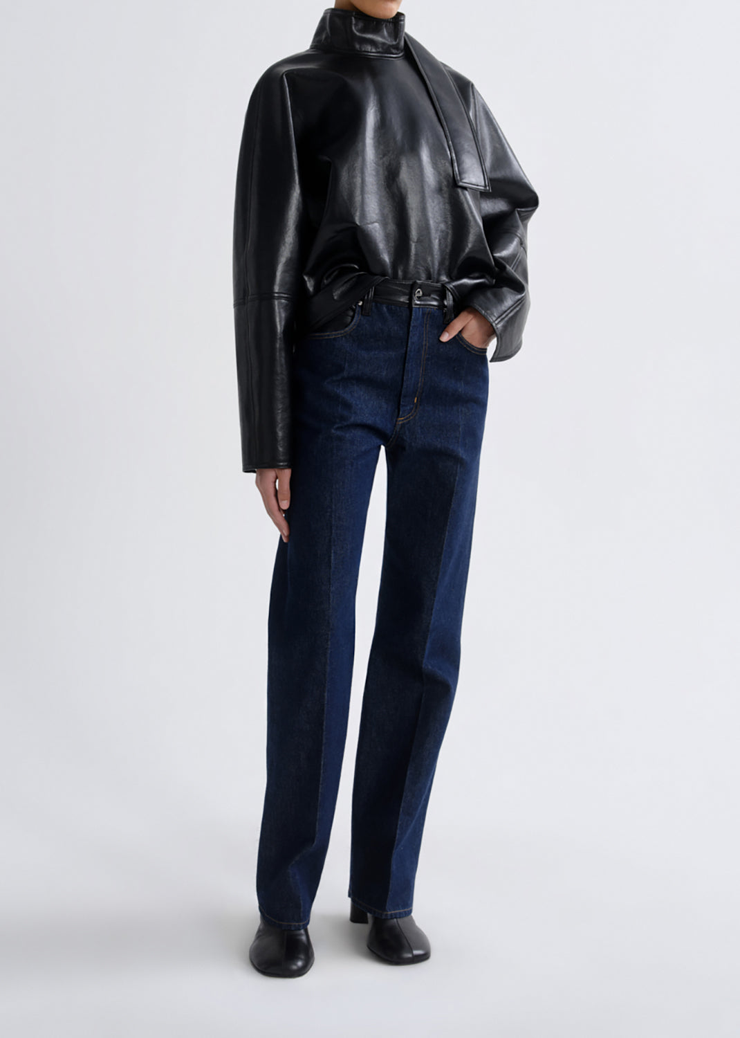 A model wearing a black leather mock-neck jacket and dark blue straight-leg jeans, standing against a grey background.