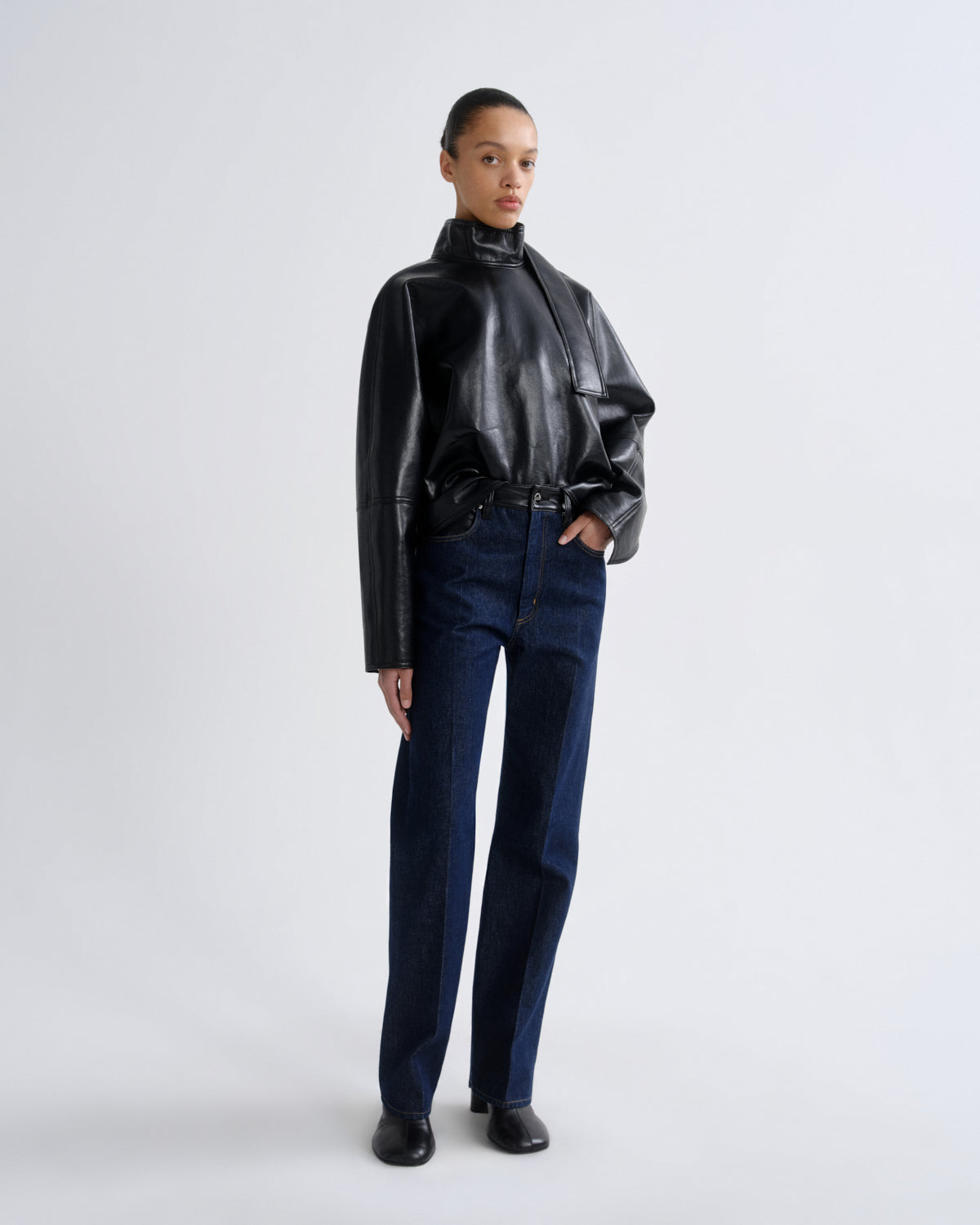 A model stands against a light gray studio background wearing a black faux-leather high-neck jacket with structured shoulders and dark blue straight-leg jeans.