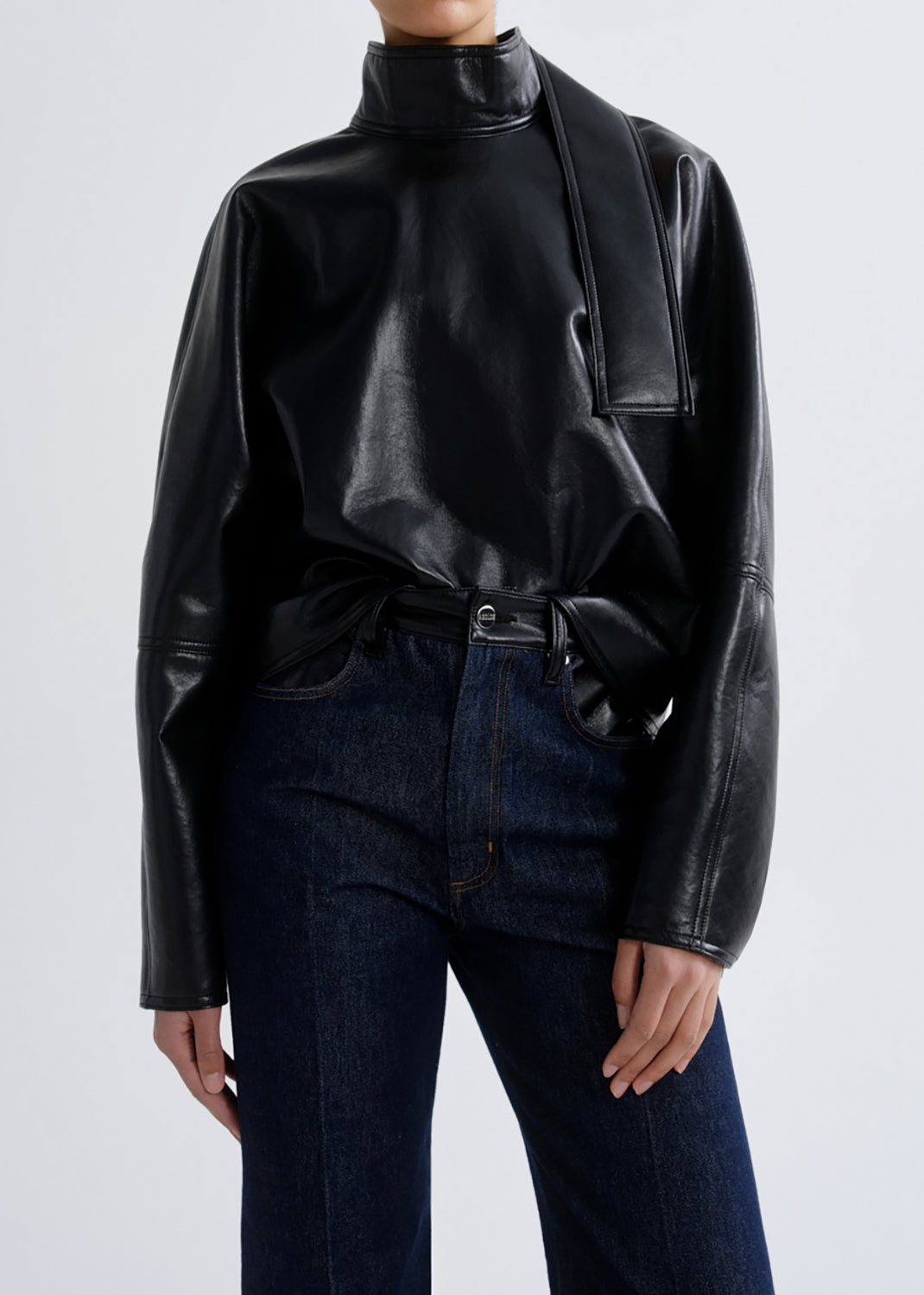 A model wearing a black faux-leather mock neck top with draped side details and dark blue jeans, shown from the neck down against a grey background.