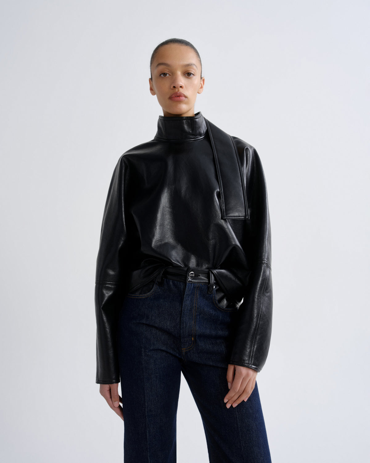 A model wearing a black faux-leather mock-neck jacket with draped sleeves and dark blue jeans, standing against a light gray studio background.