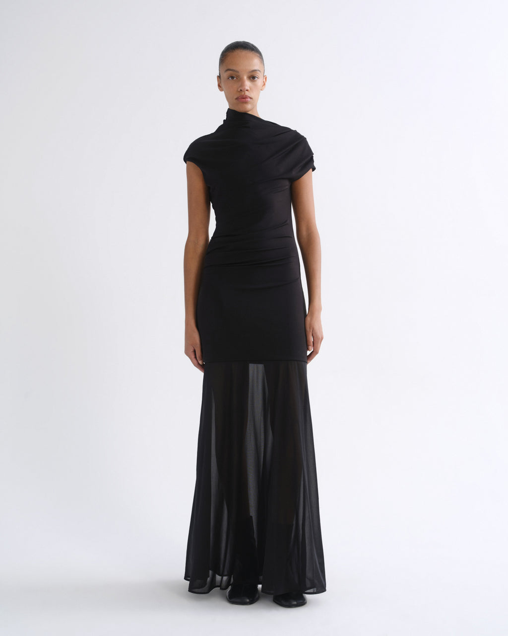 A model wearing a black high-neck, short-sleeved dress with a sheer, flowing skirt overlay, standing against a plain white studio background.