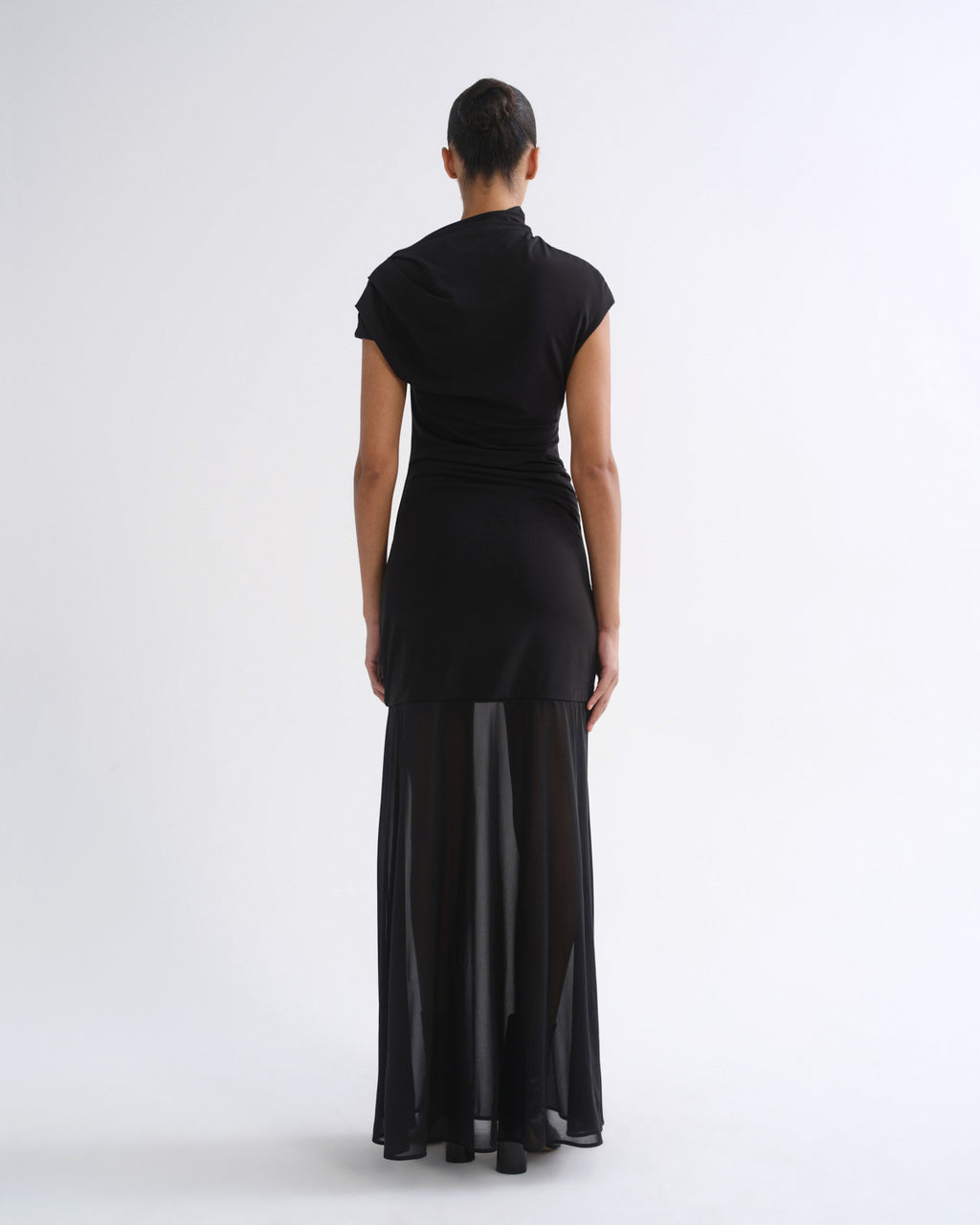 A model viewed from the back wearing a black short-sleeve mock-neck dress with a sheer black midi skirt overlay, set against a plain white studio background.