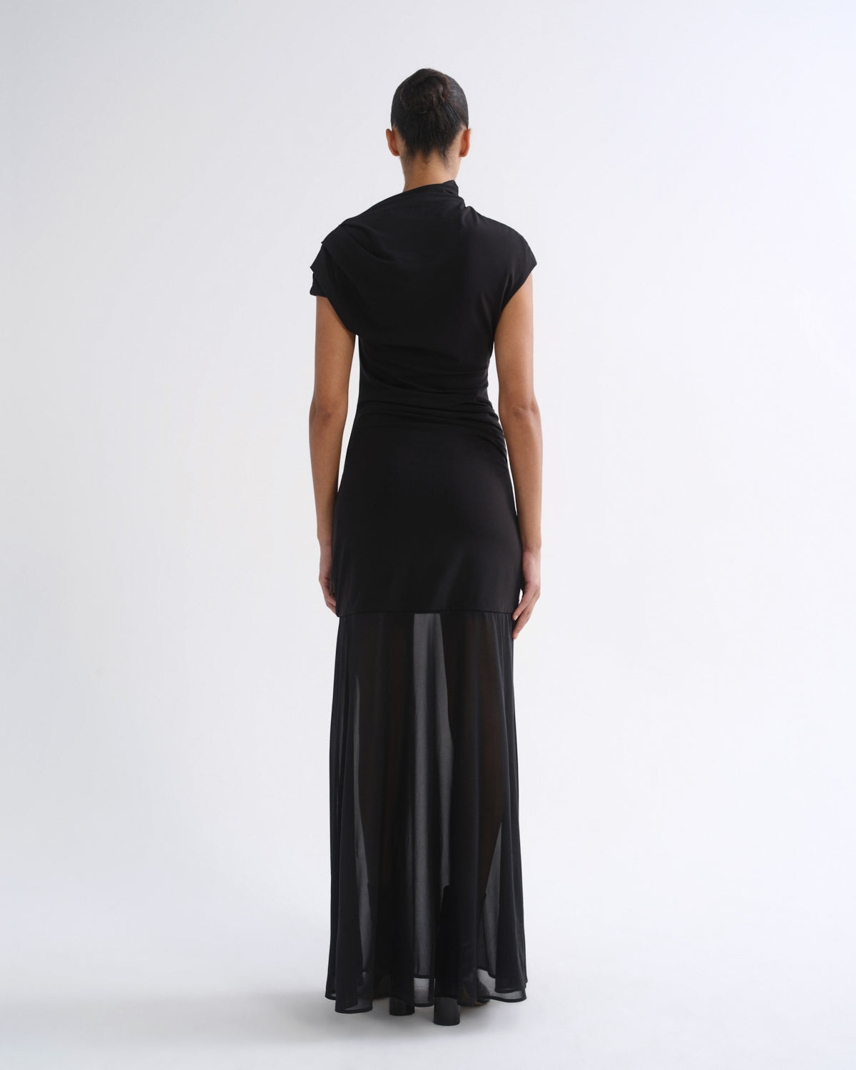 A model viewed from the back wearing a black short-sleeve mock-neck dress with a sheer black midi skirt overlay, set against a plain white studio background.