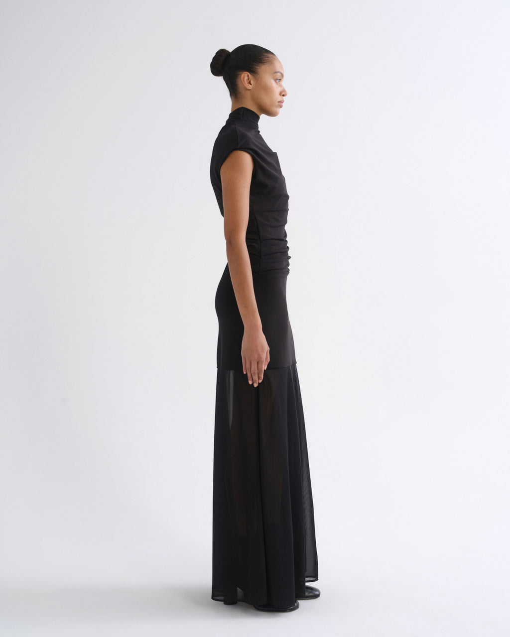 A model in profile wearing a black sleeveless high-neck maxi dress with a gathered, ruched bodice and a flowing sheer skirt, photographed against a seamless light gray studio background.
