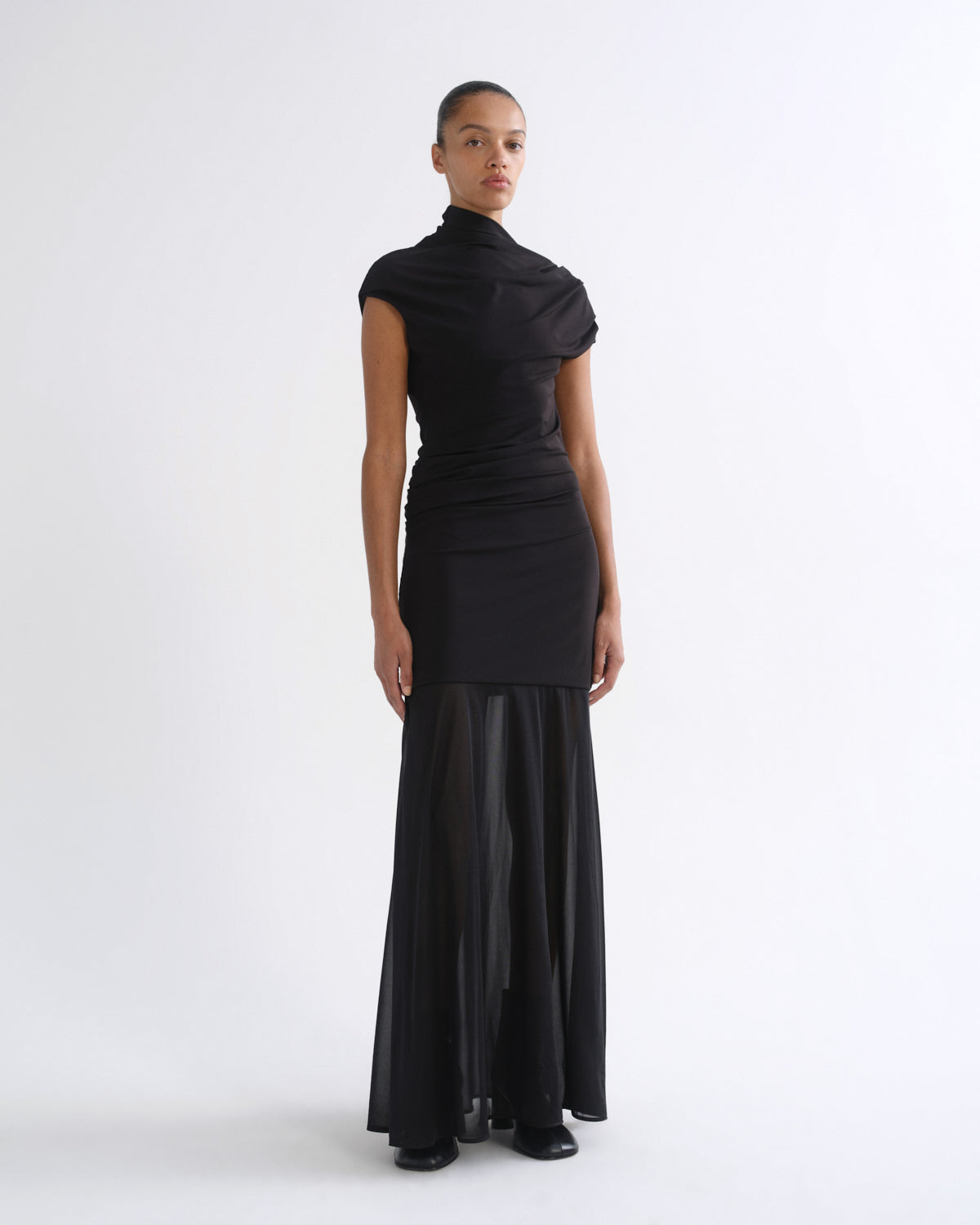 A model wearing a black high-neck, short-sleeved dress with a draped, asymmetrical bodice and a sheer, flowing skirt, photographed against a plain white studio background.