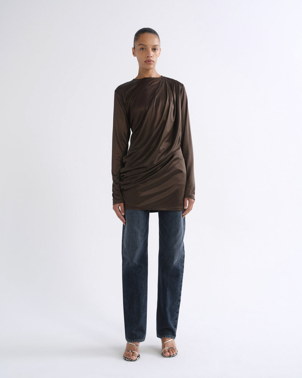 A model stands against a plain white studio background wearing a dark brown long-sleeve draped top, dark blue jeans, and flat sandals.
