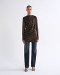 A model stands against a plain white studio background wearing a dark brown long-sleeve draped top, dark blue jeans, and flat sandals.