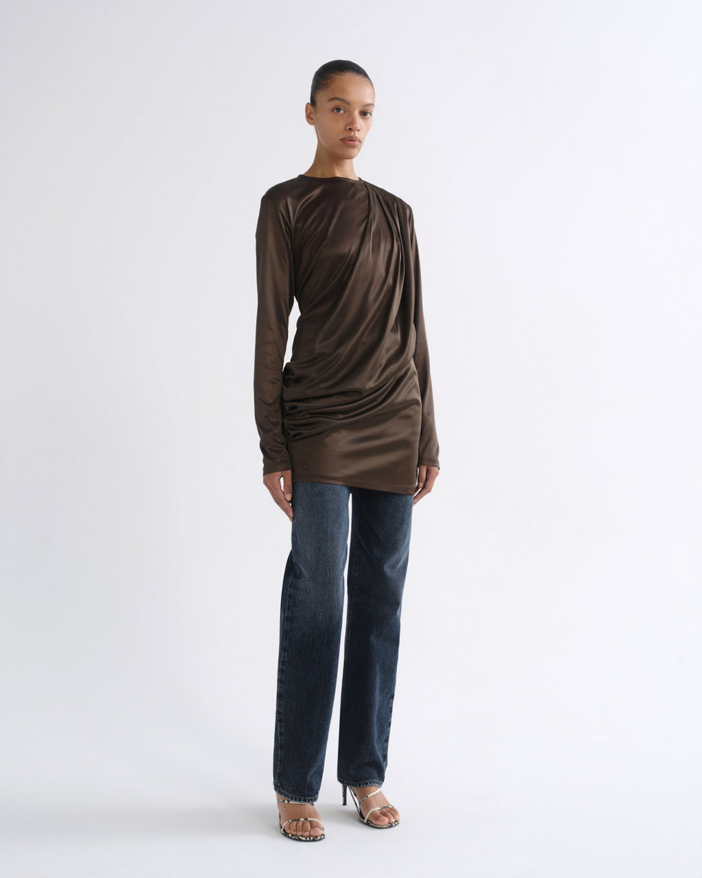 A model stands against a plain light gray studio background wearing a draped, long-sleeve satin-like top in dark brown, paired with dark blue jeans and metallic heeled sandals.