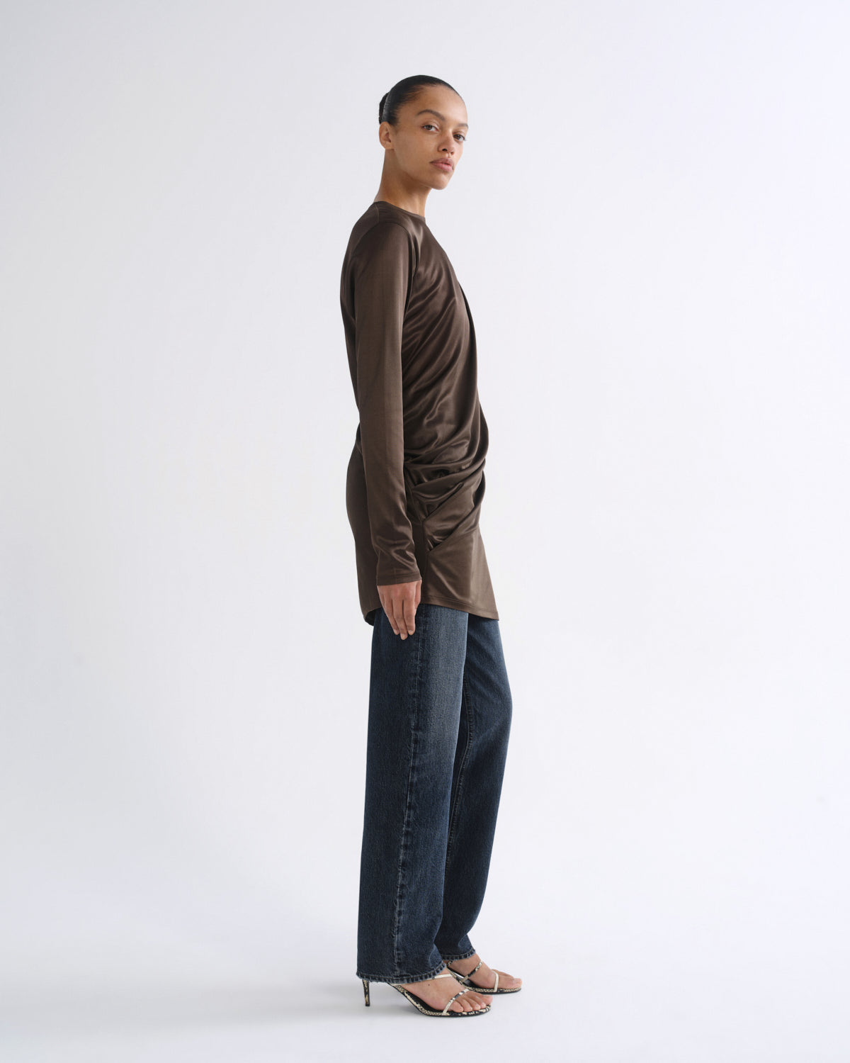 A model stands in profile wearing a draped, long-sleeve brown top, dark blue wide-leg jeans, and heeled sandals against a plain white studio background.