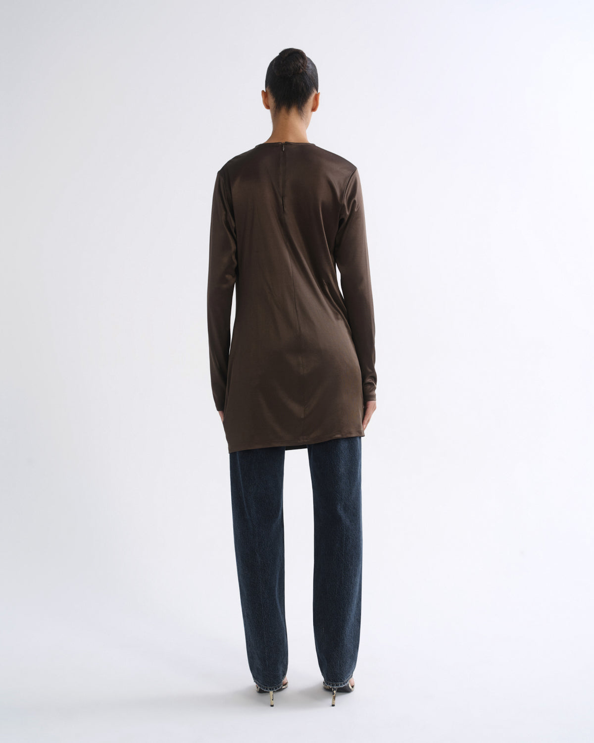 A model viewed from the back wearing a dark brown long-sleeve tunic top and dark blue jeans against a plain white studio background.