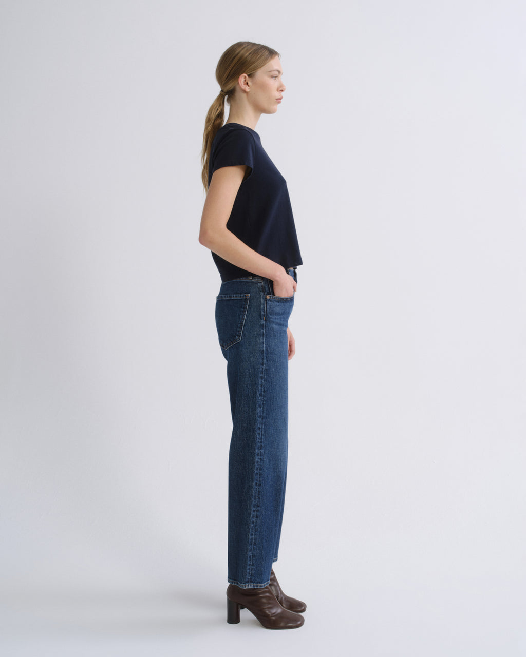 A woman in side profile wearing a dark navy short-sleeve top, blue straight-leg jeans, and brown heeled ankle boots, standing against a light gray studio background.