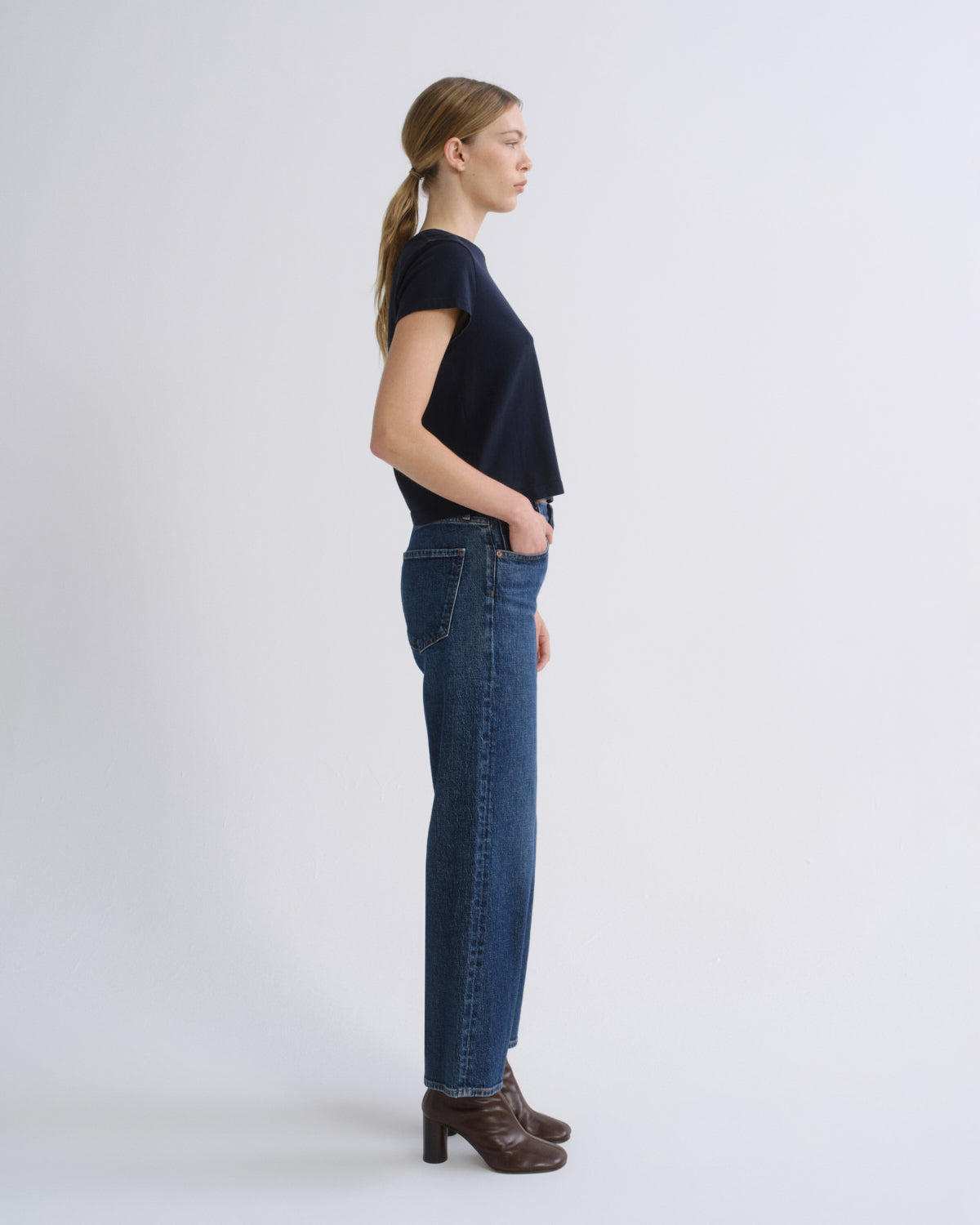 A woman in side profile wearing a dark navy short-sleeve top, blue straight-leg jeans, and brown heeled ankle boots, standing against a light gray studio background.