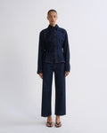 A model stands against a light gray studio background wearing a dark indigo denim two-piece set consisting of a cropped long-sleeve button-up shirt and matching wide-leg cropped pants.