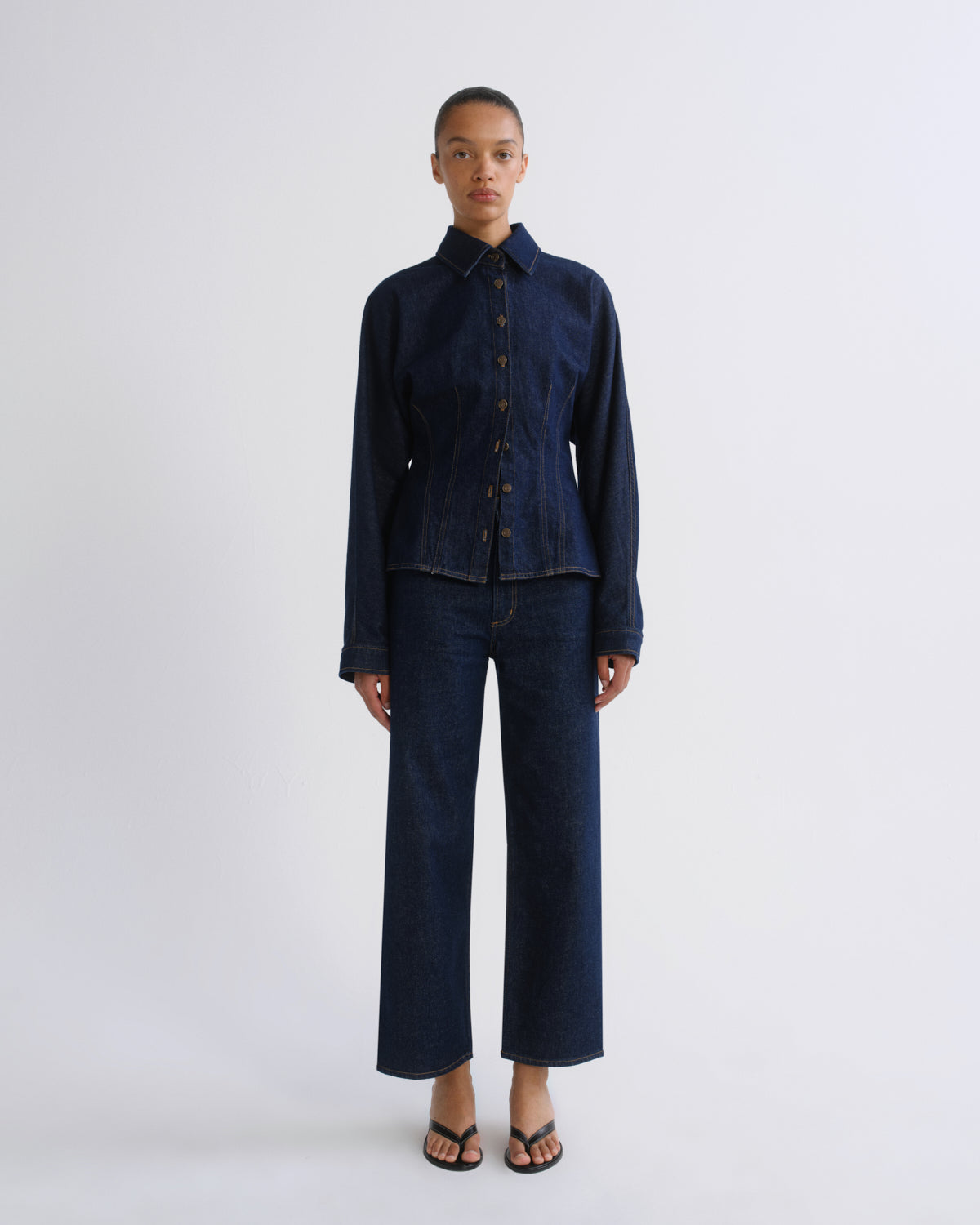 A model stands against a light gray studio background wearing a dark indigo denim two-piece set consisting of a cropped long-sleeve button-up shirt and matching wide-leg cropped pants.