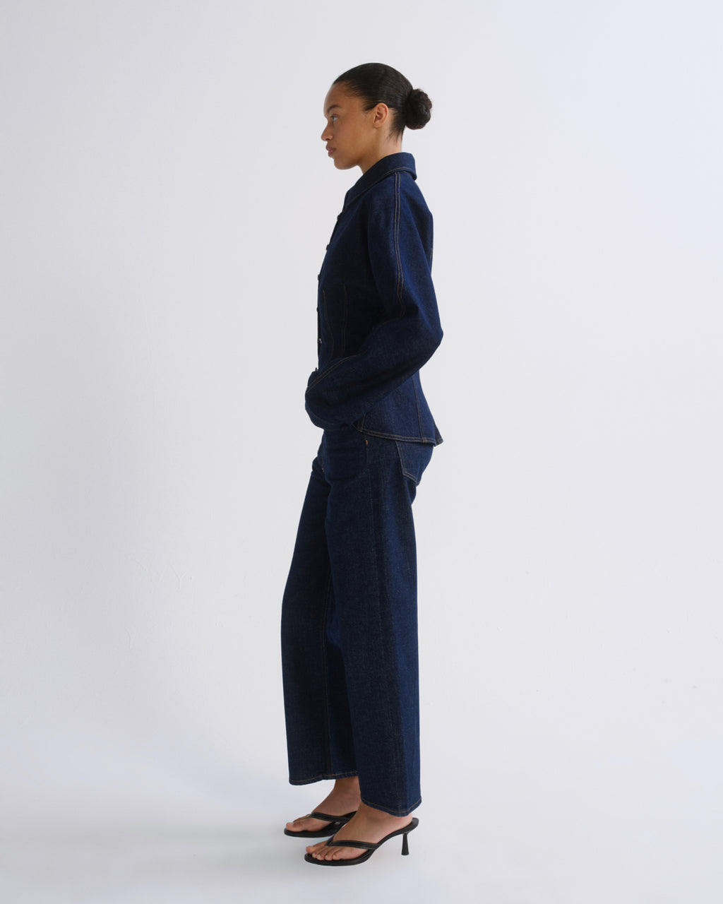 A model in profile wearing a dark indigo denim two-piece set consisting of a long-sleeve button-up shirt and matching wide-leg trousers, paired with black heeled sandals, against a plain light gray studio background.