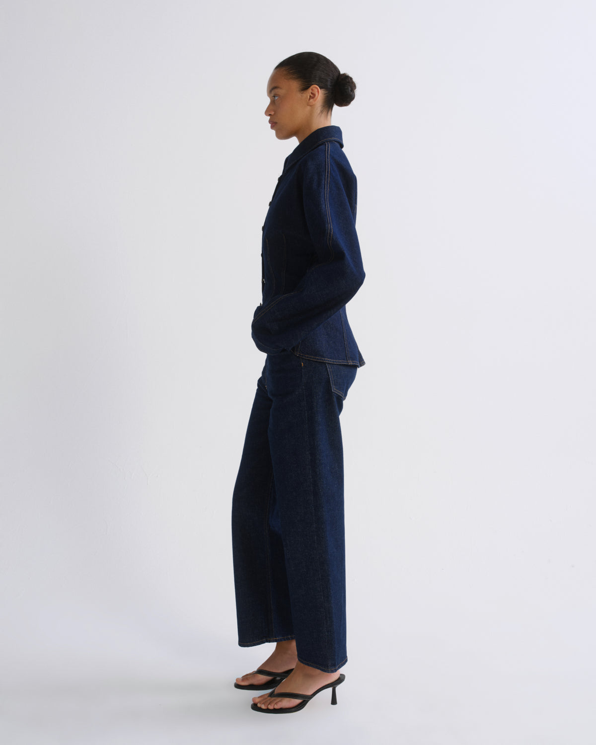 A model in profile wearing a dark indigo denim two-piece set consisting of a long-sleeve button-up shirt and matching wide-leg trousers, paired with black heeled sandals, against a plain light gray studio background.