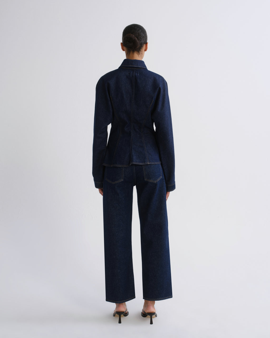 A model viewed from the back wearing a dark indigo denim two-piece set consisting of a long-sleeve collared jacket and matching wide-leg trousers, paired with black high heels against a light gray studio background.