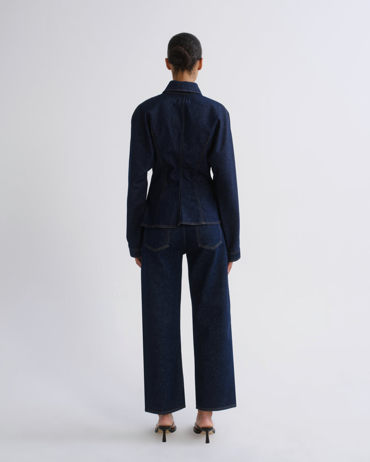 A model viewed from the back wearing a dark indigo denim two-piece set consisting of a long-sleeve collared jacket and matching wide-leg trousers, paired with black high heels against a light gray studio background.