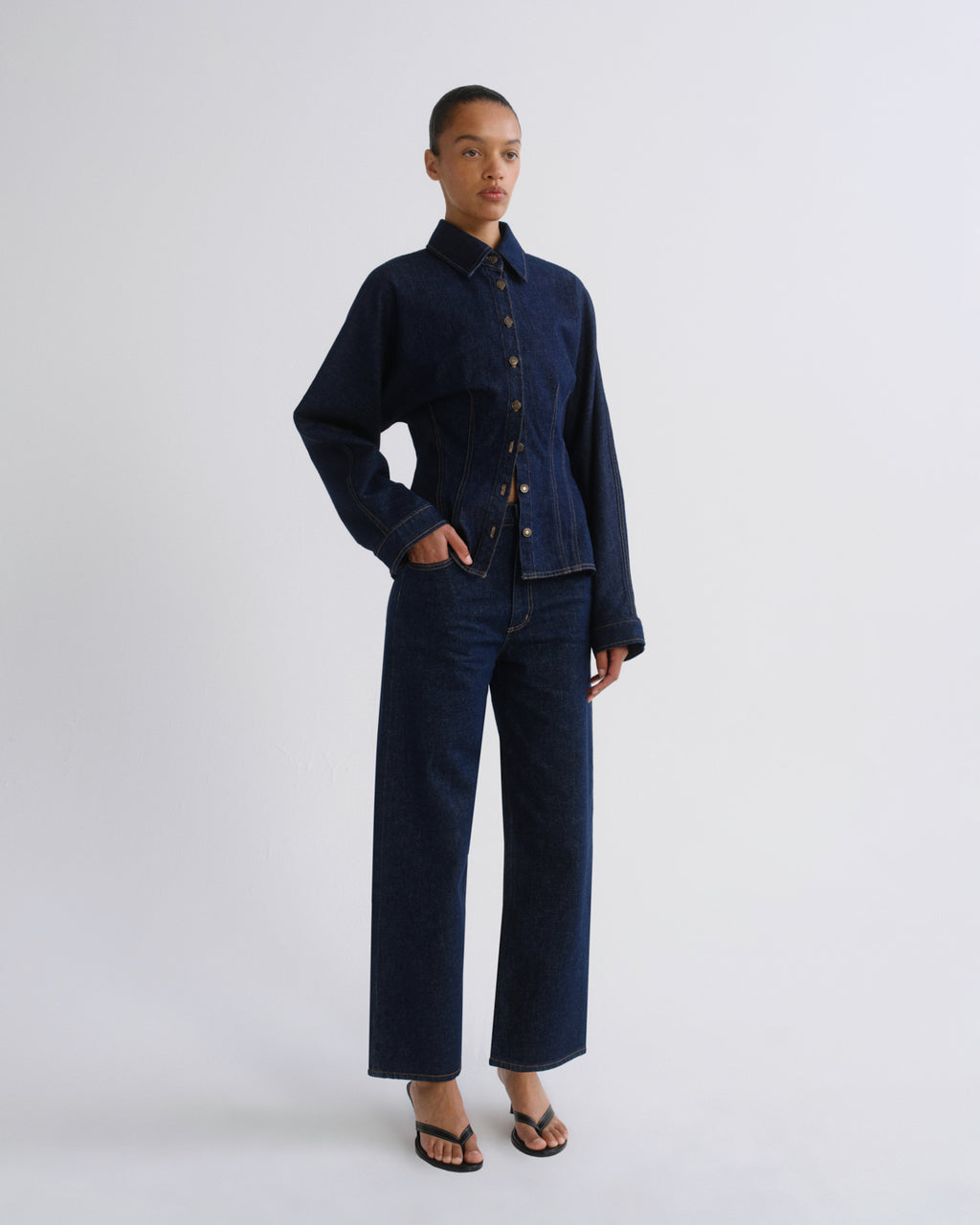 A model stands against a light gray studio background wearing a dark indigo denim two-piece set consisting of a cropped button-front jacket and matching wide-leg pants, paired with black sandals.