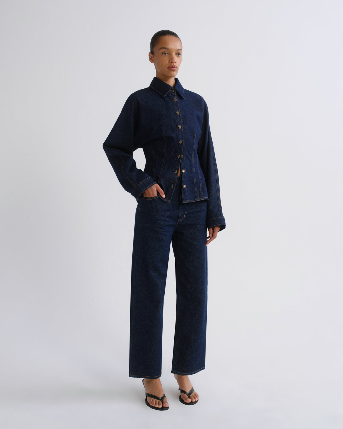 A model stands against a light gray studio background wearing a dark indigo denim two-piece set consisting of a cropped button-front jacket and matching wide-leg pants, paired with black sandals.
