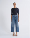 A model stands against a light gray studio background wearing a dark navy three-quarter sleeve top, blue wide-leg cropped jeans, and black sandals.