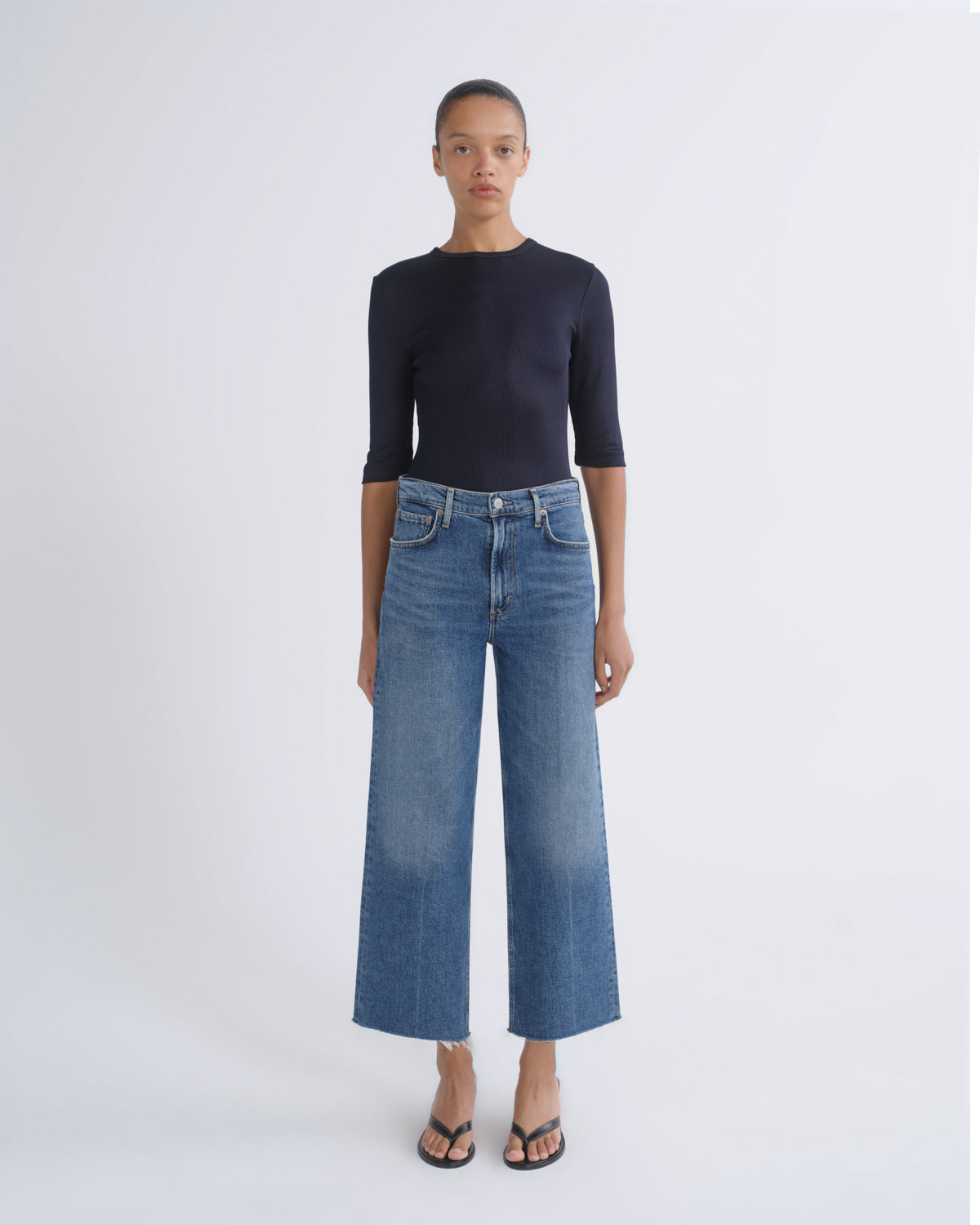 A model stands against a light gray studio background wearing a dark navy three-quarter sleeve top, blue wide-leg cropped jeans, and black sandals.