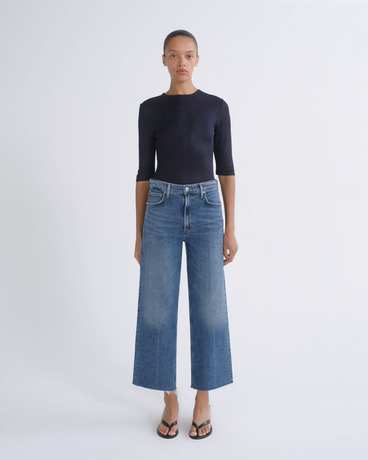 A model stands against a light gray studio background wearing a dark navy three-quarter sleeve top, blue wide-leg cropped jeans, and black sandals.