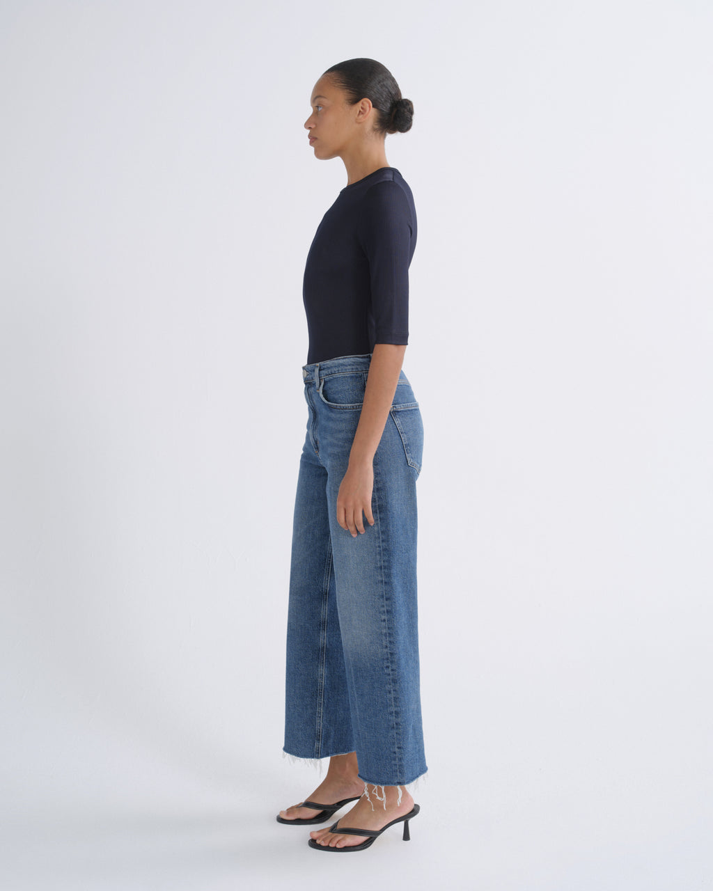 A woman in profile wearing a dark navy short-sleeve top, blue wide-leg cropped jeans, and black heeled sandals, standing against a plain light gray studio background.