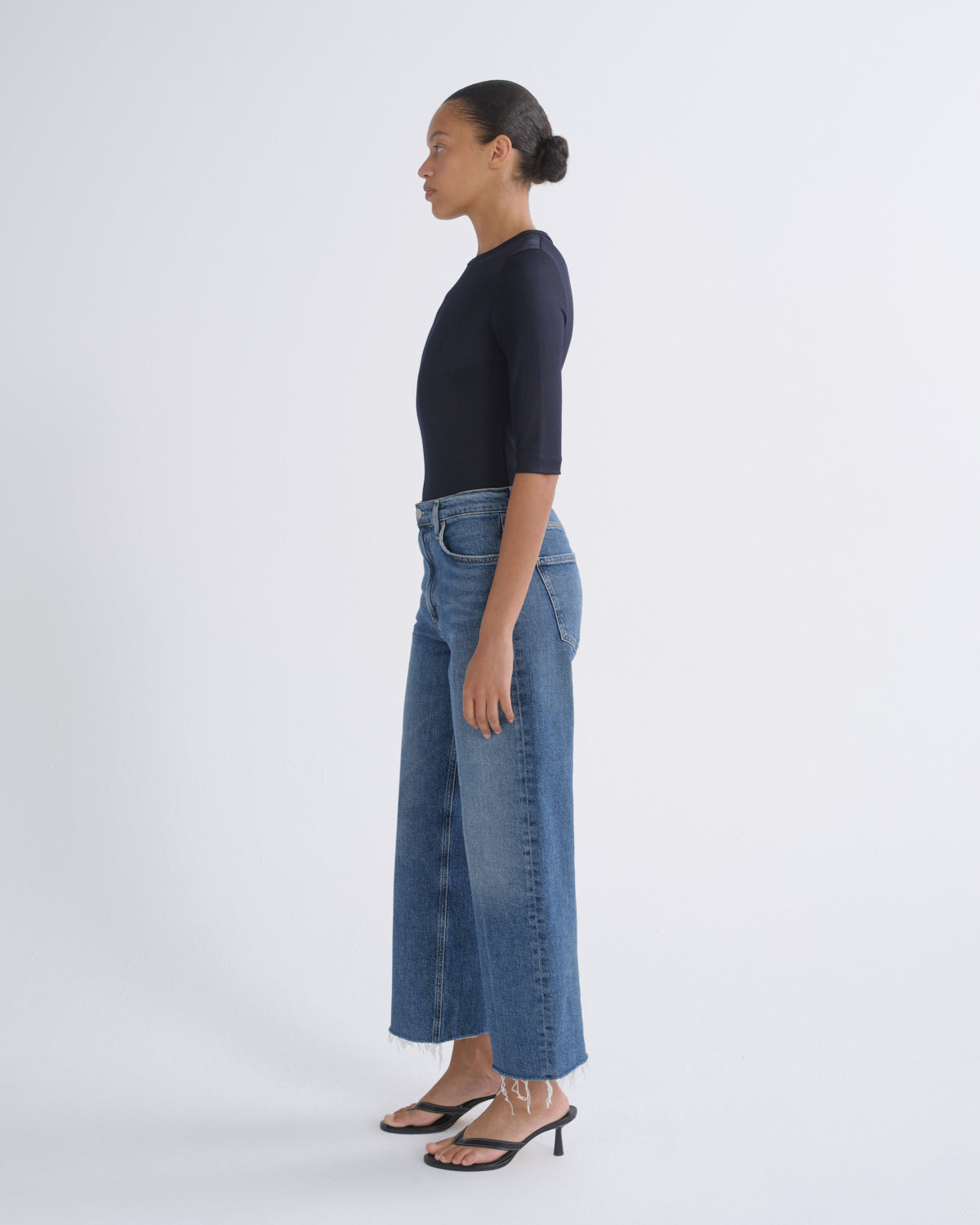 A woman in profile wearing a dark navy short-sleeve top, blue wide-leg cropped jeans, and black heeled sandals, standing against a plain light gray studio background.