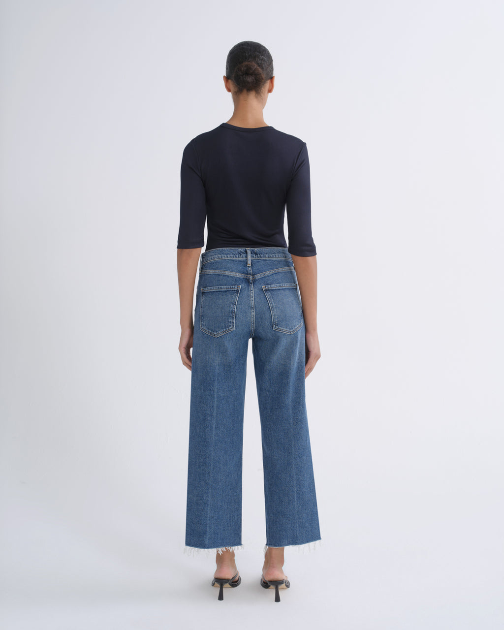 A model viewed from the back wearing a dark navy three-quarter sleeve top, blue wide-leg cropped jeans, and black high heels against a plain light gray studio background.