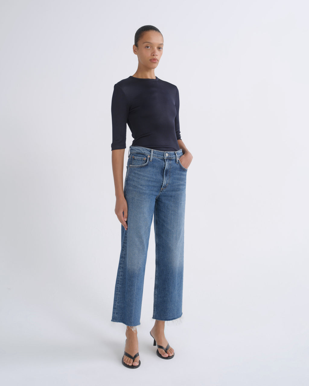 A model stands against a light gray studio background wearing a dark navy long-sleeve top, blue cropped wide-leg jeans, and black flip-flop sandals.