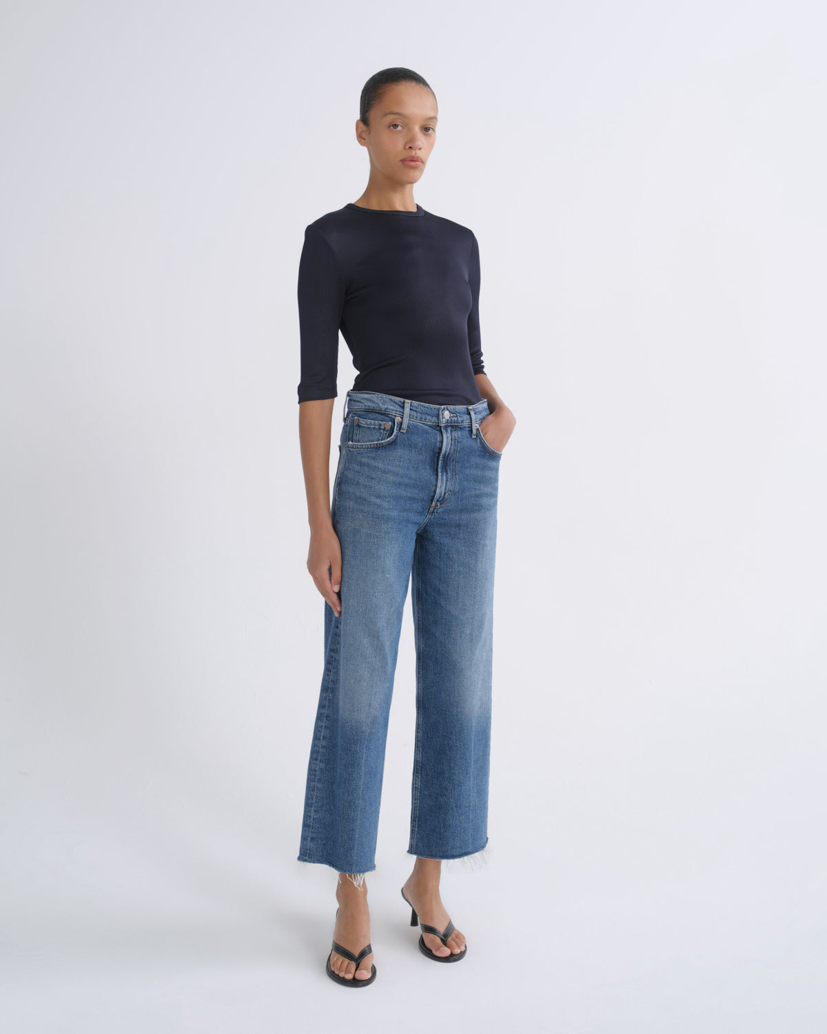 A model stands against a light gray studio background wearing a dark navy long-sleeve top, blue cropped wide-leg jeans, and black flip-flop sandals.