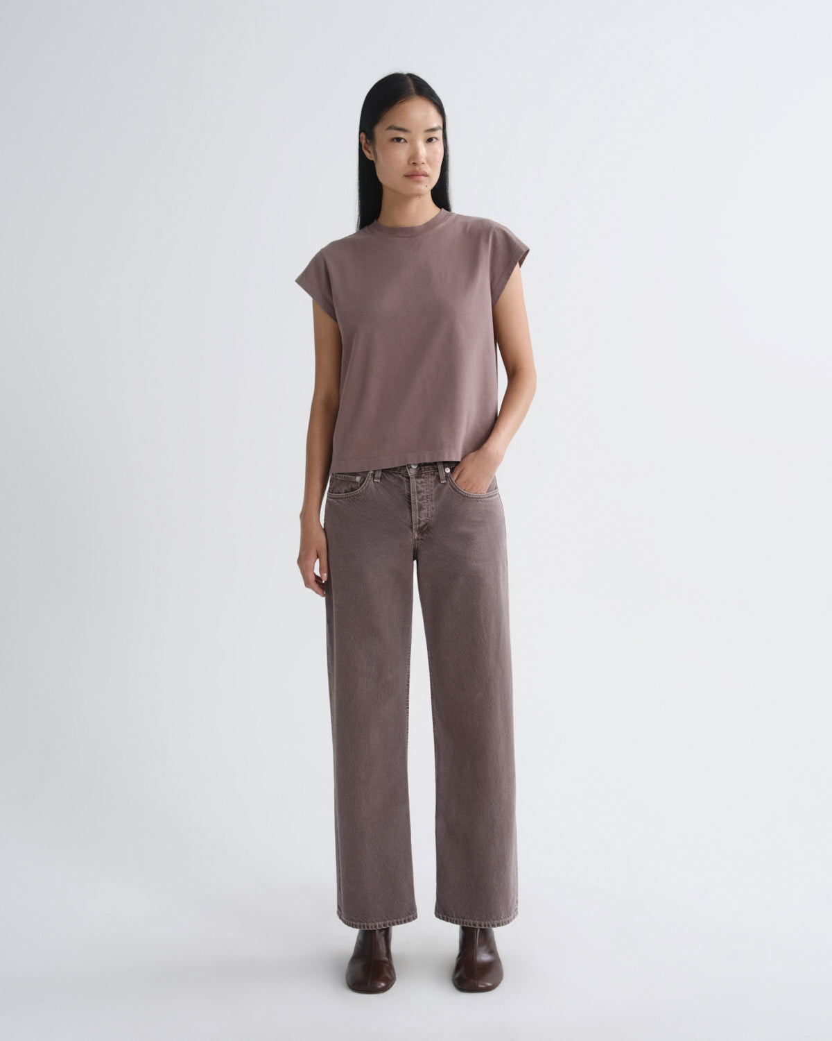 A model stands against a light gray studio background wearing a muted mauve-brown short-sleeve top and matching wide-leg pants, with dark brown boots.