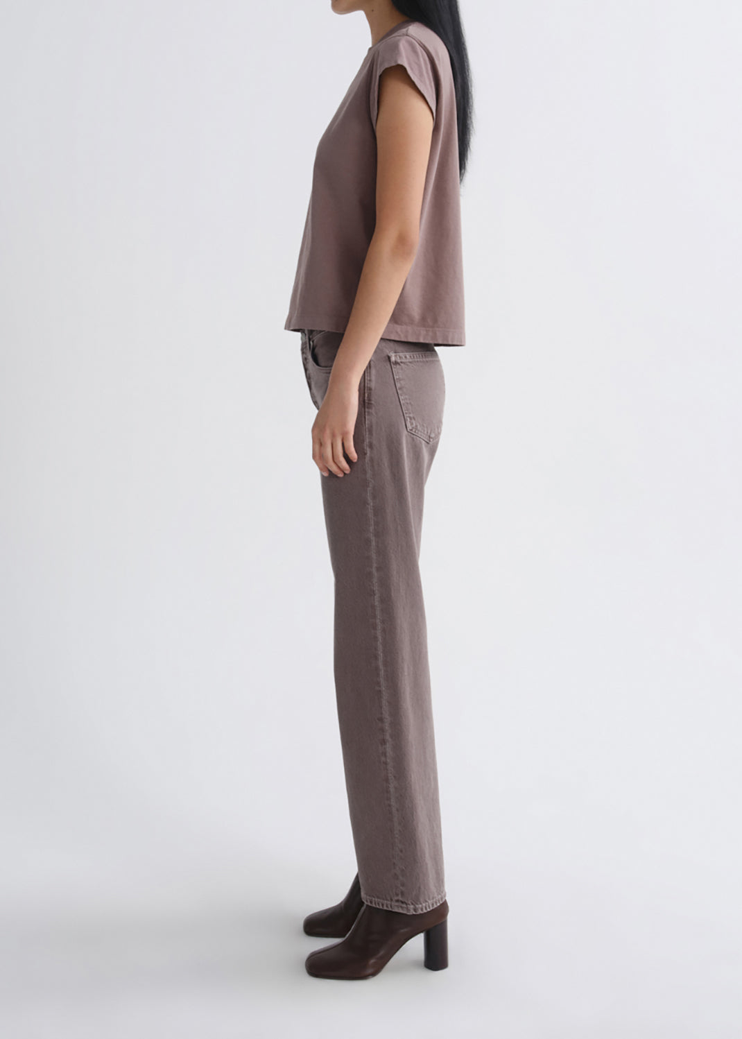 A side-profile studio shot of a model wearing a muted mauve-brown sleeveless top, matching wide-leg jeans, and dark brown block-heel ankle boots against a grey background.