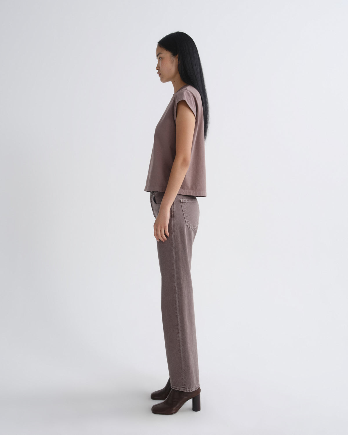 A woman stands in profile wearing a sleeveless taupe top and matching straight-leg jeans with dark brown heeled ankle boots, photographed against a clean light gray studio background.