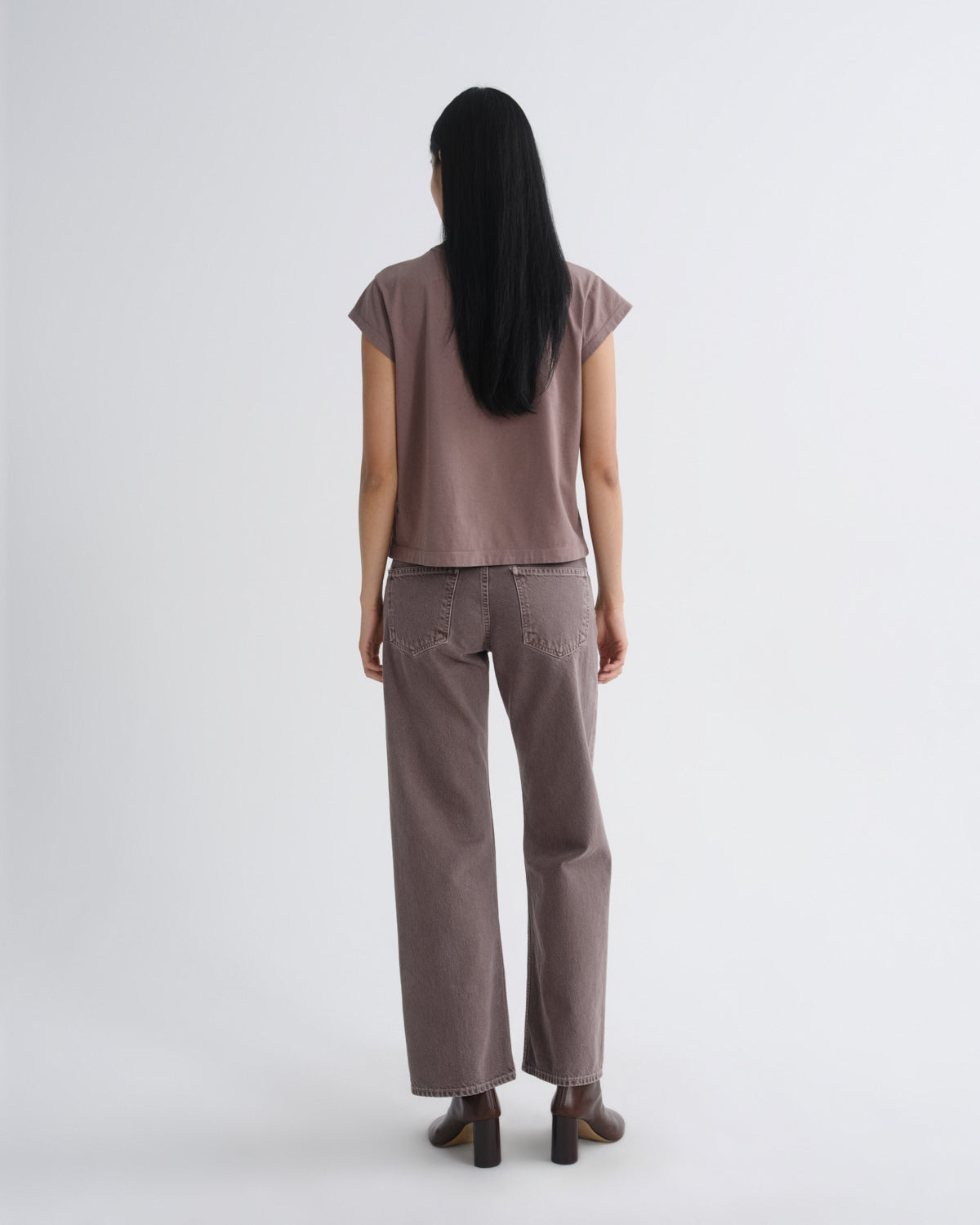 A woman viewed from behind wearing a mauve-brown short-sleeve cropped t-shirt and matching wide-leg trousers, paired with dark brown block-heel boots, against a plain light gray studio background.