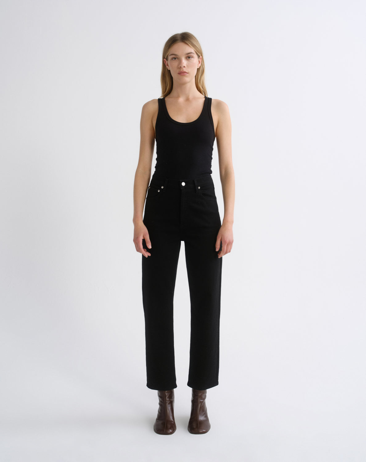 A model stands against a plain light gray studio background wearing a black sleeveless top, black high-waisted straight-leg jeans, and dark brown ankle boots.