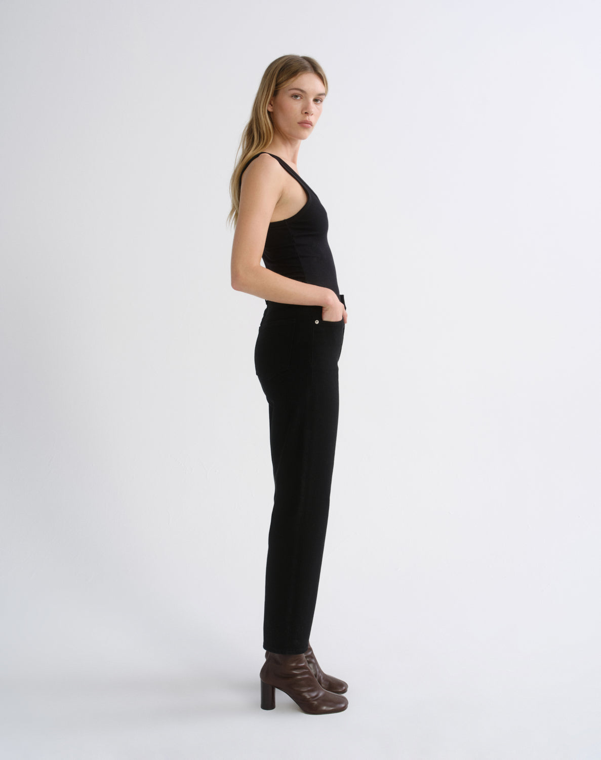 A woman stands in profile wearing a black sleeveless top and black slim-fit pants, paired with dark brown heeled ankle boots, against a plain light gray studio background.