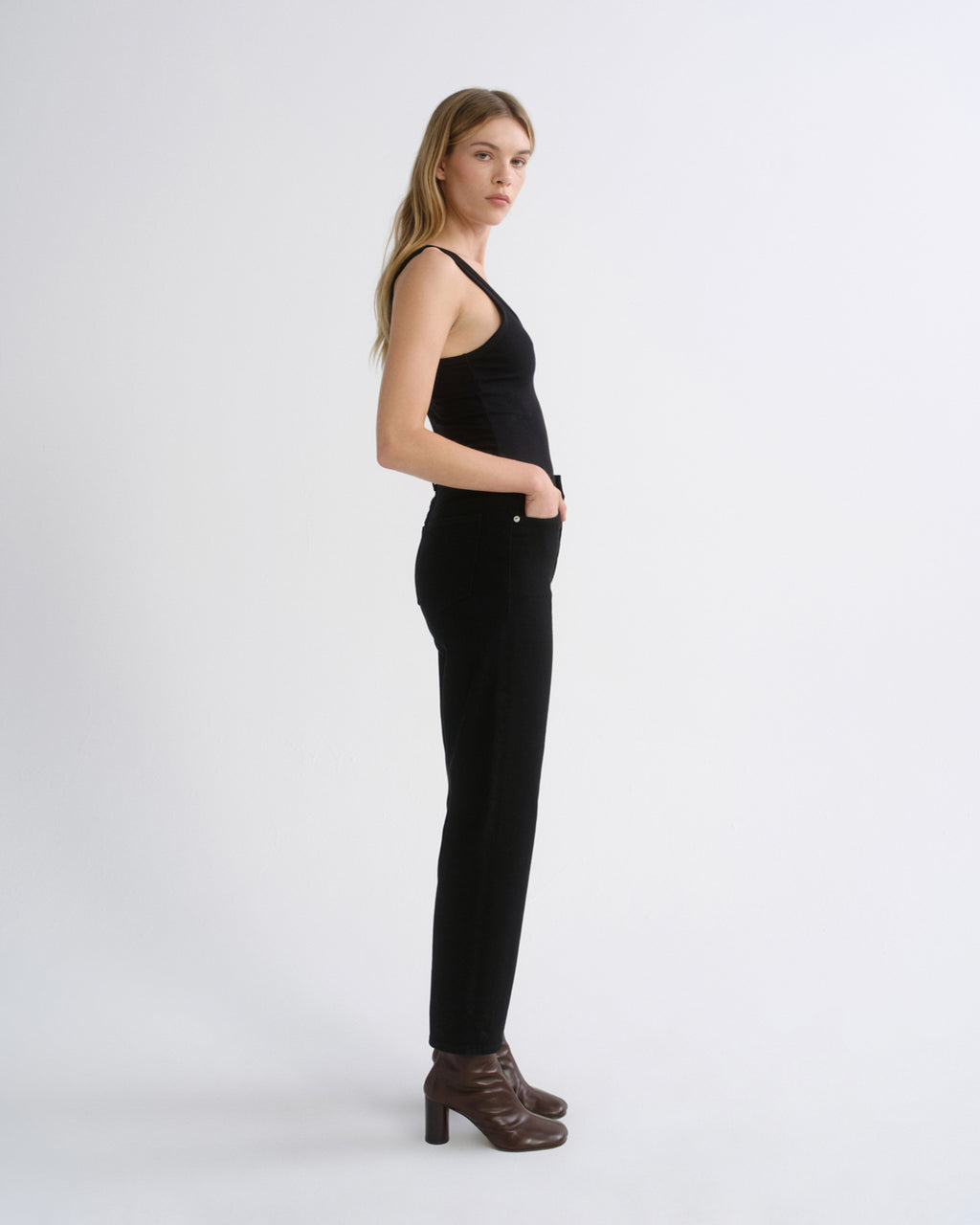 A woman stands in profile wearing a black sleeveless top and black slim-fit pants, paired with dark brown heeled ankle boots, against a plain light gray studio background.