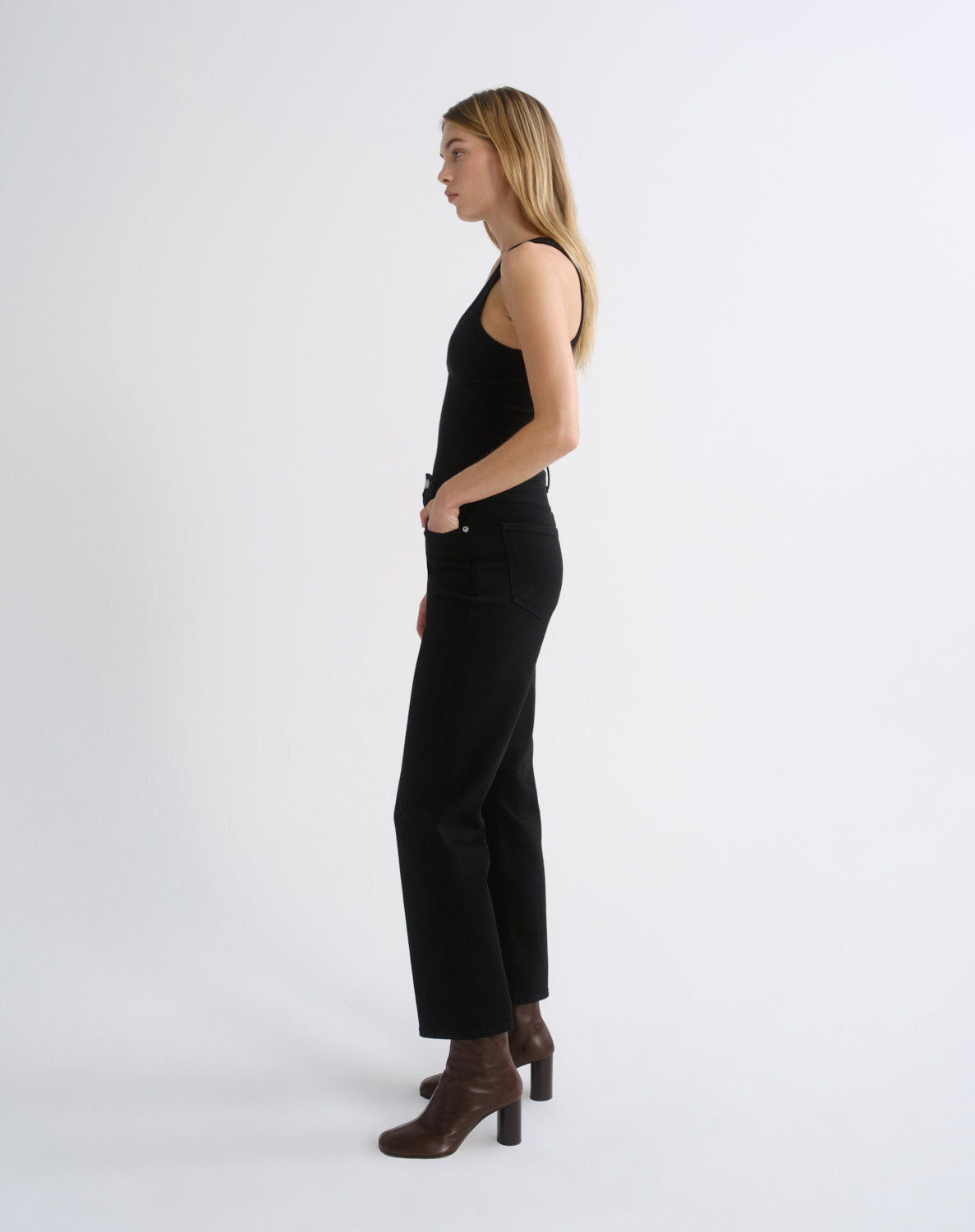 A woman stands in profile wearing a black sleeveless top and black wide-leg pants with brown heeled ankle boots, photographed against a clean light gray studio background.