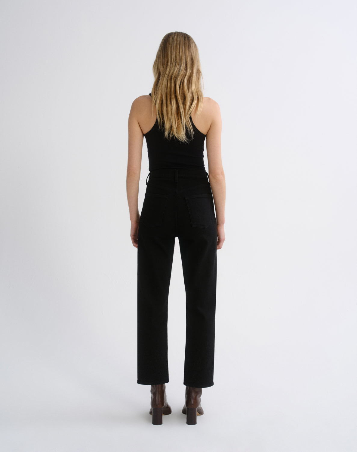 A woman viewed from the back wearing a black sleeveless top and black wide-leg cropped pants, standing against a plain light gray studio background.