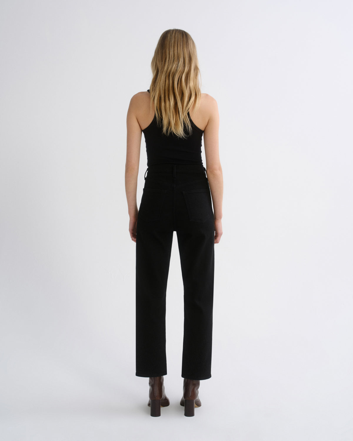 A woman viewed from the back wearing a black sleeveless top and black wide-leg cropped pants, standing against a plain light gray studio background.